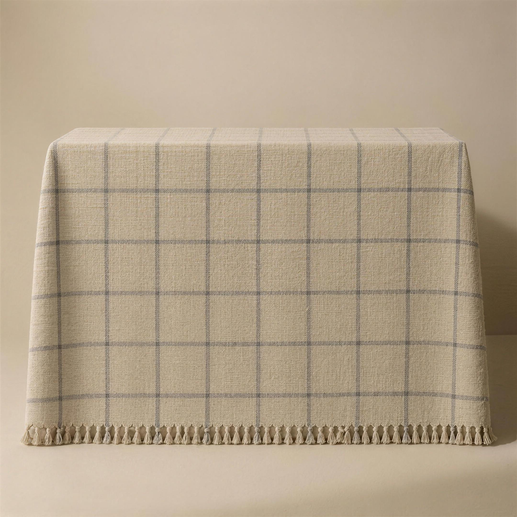 The Woven in bone, draped face-up over a plinth, the windowpane grid woven into the cloth in grey and a knotted fringe along the lower edge.