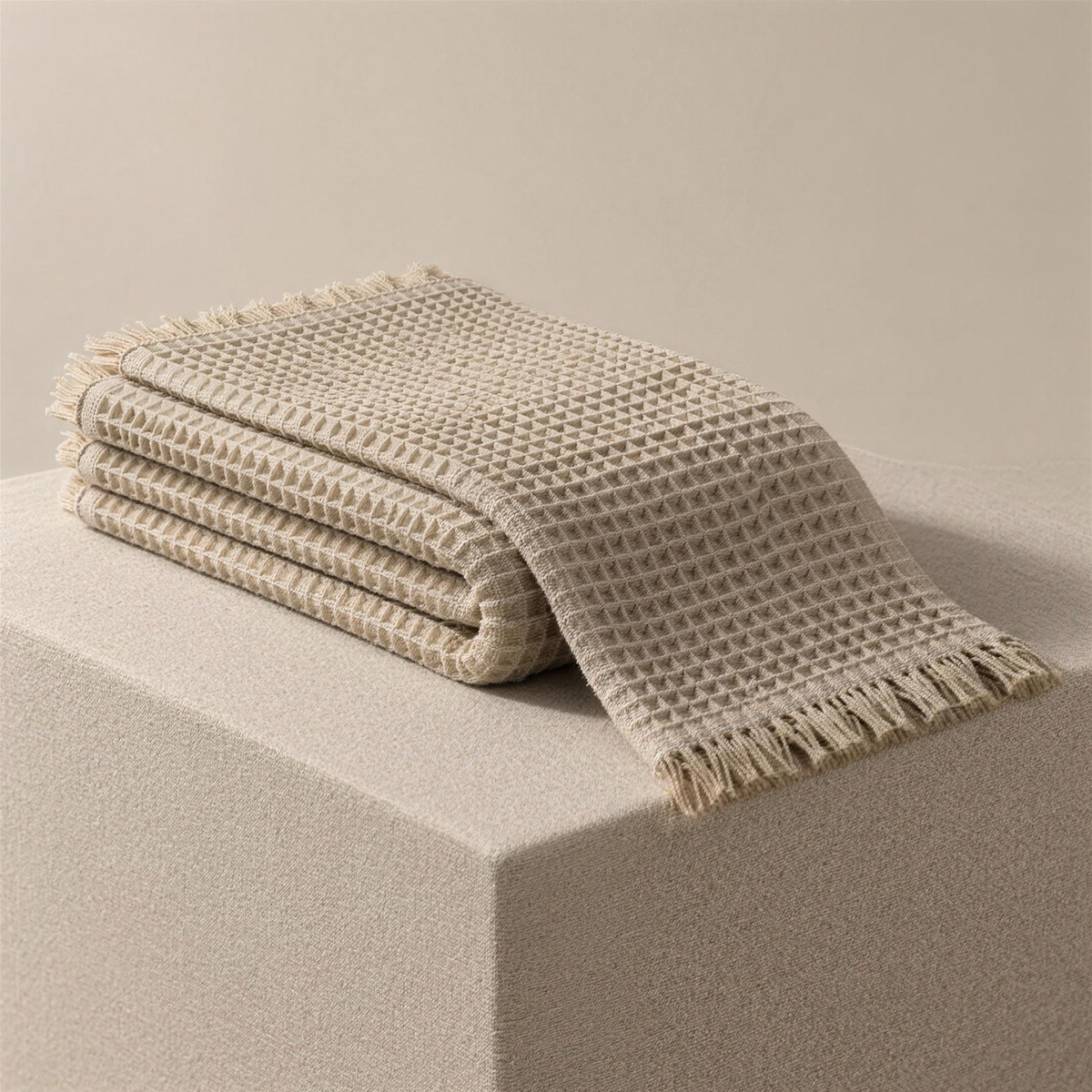 The Waffle in bone, folded on a plinth with the top layer fallen open, showing the deep waffle pockets and the fringed end.