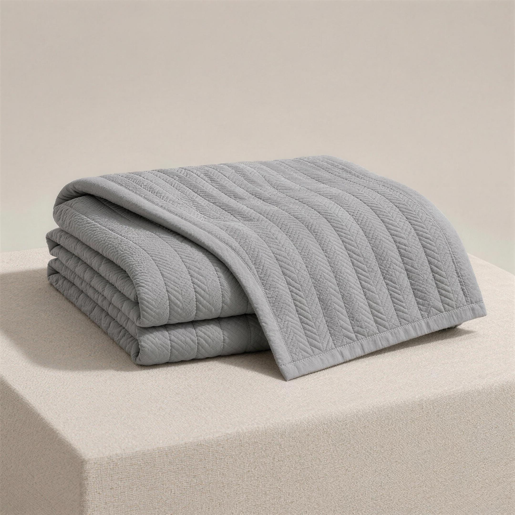 The Quilt in ash, folded on a plinth, narrow chevron-stitched columns catching the light.
